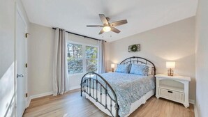 Une chambre à coucher comprenant un lit, un ventilateur de plafond, une table de chevet, une fenêtre avec des rideaux et une vue sur les arbres à l’extérieur.