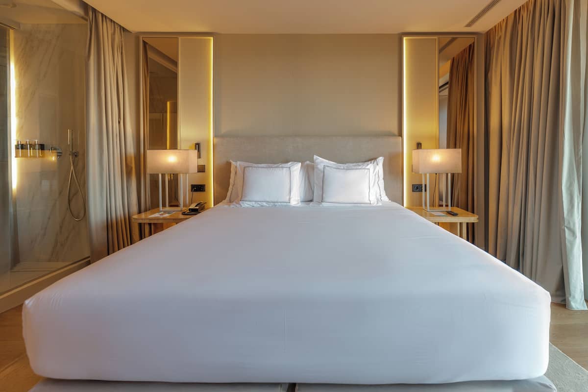 A large bed with white linens, two bedside tables with lamps, a mirror, and a shower area.
