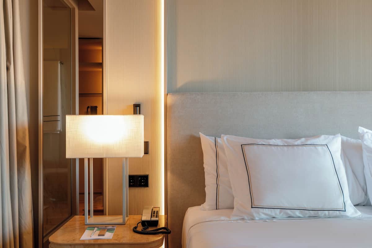A hotel room with a bed, a bedside table with a lamp, a phone, and a mirror.