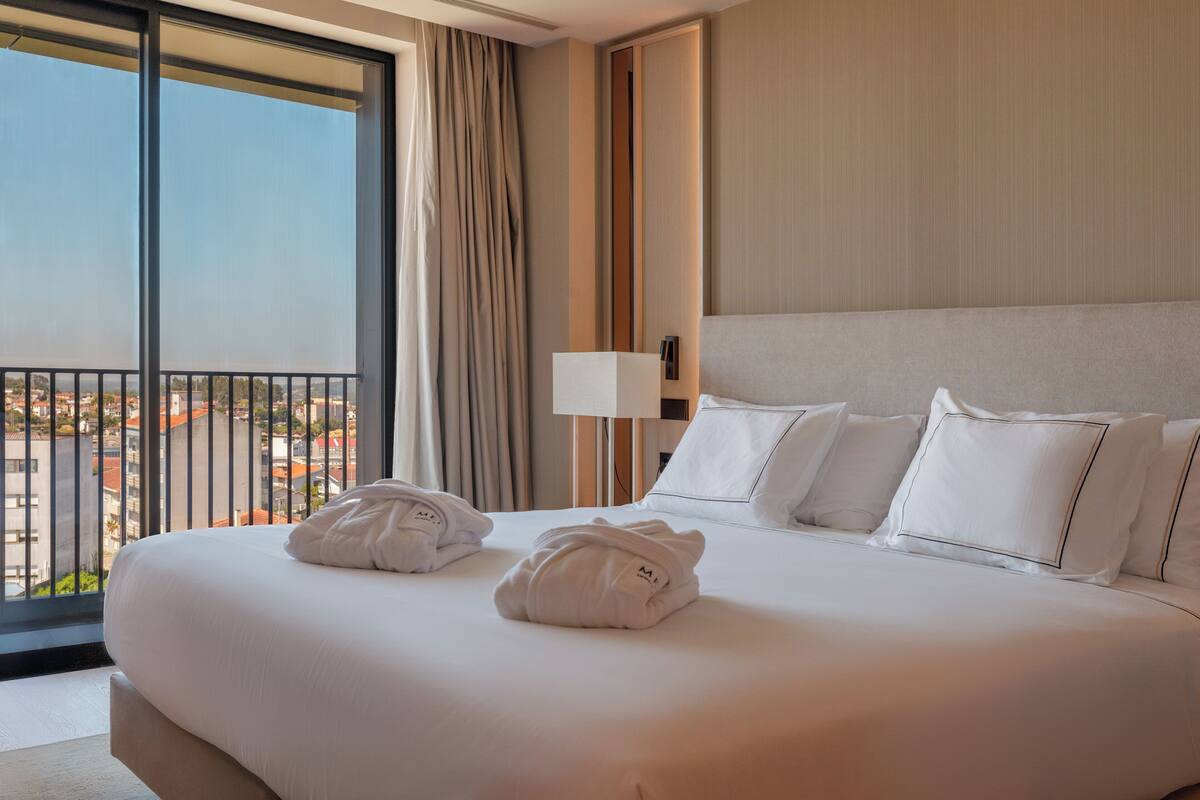 A hotel room with a large bed, two towels, a nightstand with a lamp, and a view of the city through a balcony.
