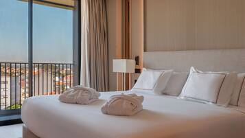 Quarto de hotel com uma cama grande, duas toalhas, uma mesa de cabeceira com abajur e vista para a cidade através de uma varanda.