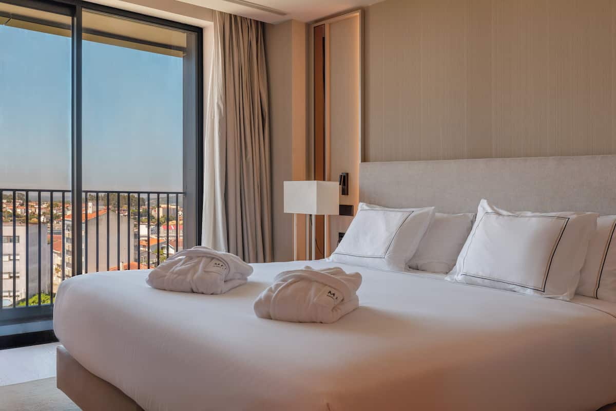 A hotel room with a large bed, a nightstand, a lamp, and a view of the city through a balcony.