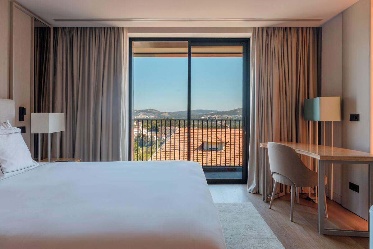 A hotel room with a bed, a desk, a chair, a large window with curtains, and a view of the cityscape.