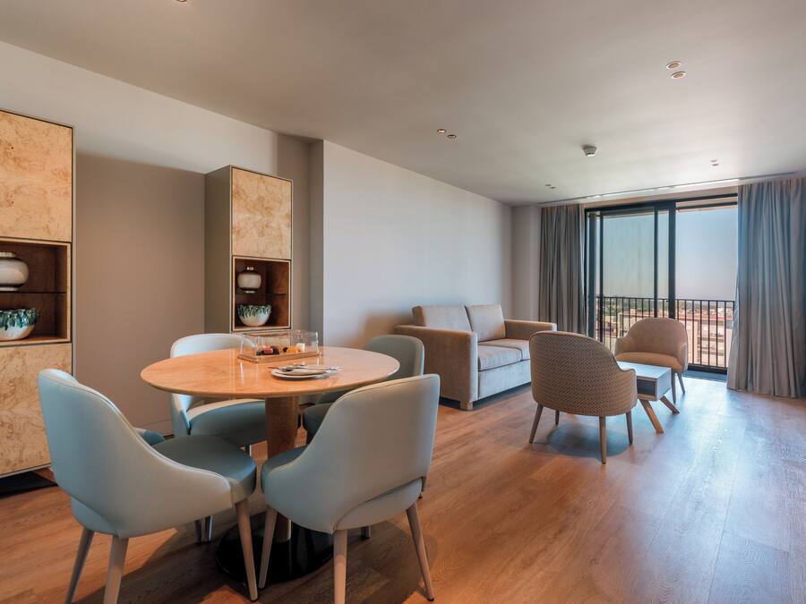 A modern living room with a round wooden table, light blue chairs, a beige sofa, and a large window with curtains.