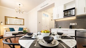 A modern dining area with a round table set for four, a white sofa with blue cushions, and a kitchen with white cabinets and a microwave. - Asty Luxury Apartments (Athens)