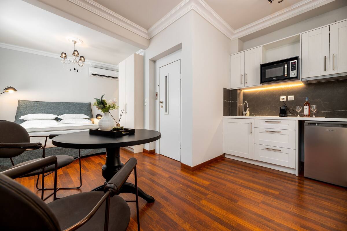 A modern hotel room with a wooden floor, a round dining table, a bed with a grey headboard, and a kitchenette with white cabinets and a microwave.