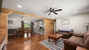 A spacious living area with a wooden floor, a ceiling fan, a brown leather sofa, a dining table with chairs, and a kitchen area in the background. - 4 bedroom house WiFi/AC/deck/firepit/grill Laurel Lake/Cumberland Falls, Corbin (Corbin)