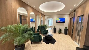 Moderni aula, jossa on puupanelointia, vihreitä nojatuoleja, suuri peili, litteänäyttöinen televisio ja ruukkukasveja.