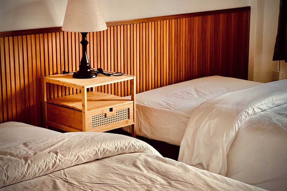 Une chambre d’hôtel avec deux lits, une tête de lit en bois, une table de chevet avec une lampe et une petite étagère en bois.