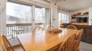 A dining area with a wooden table and chairs, a chandelier, and a view of the outdoors through large sliding glass doors. - Cozy Sugarbush condo w/ ski-in/ski-out (Warren)