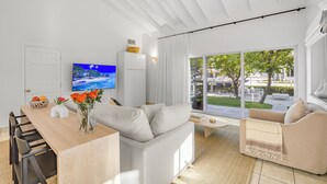 A modern living room with a wooden dining table, white sofas, a flat-screen TV, and a large window with a view of the outdoors. - Captian's Suite @ Yacht Club, Large Suite 5-minute walk from Grace Bay Beach (Providenciales)