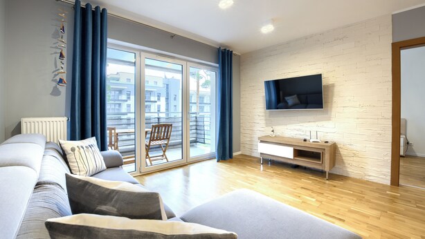 A modern living room with a sofa, a TV mounted on a brick wall, and a view of the outside through large windows. - Apartamenty Sun & Snow Blue Baltic (Pogorzelica)