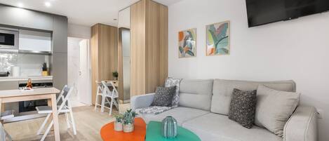 A modern living room with a sofa, two small tables, a TV mounted on the wall, and a kitchen area in the background.