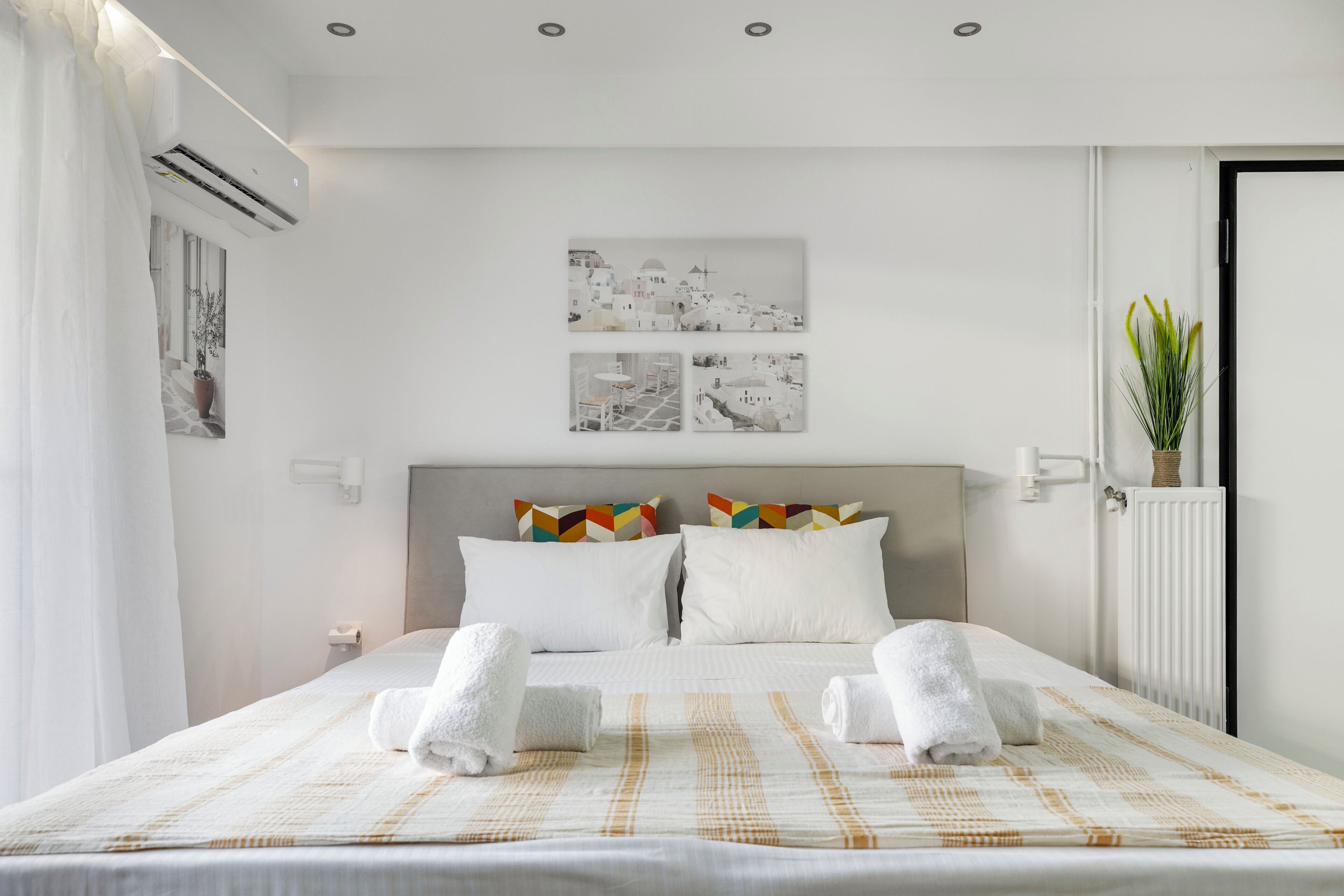 A hotel room with a large bed, white bedding, two pillows, a headboard, a wall-mounted air conditioner, a radiator, and a potted plant.