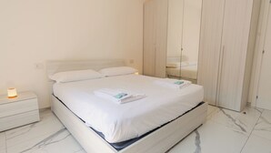 Ein ordentlich bezogenes Bett mit weißen Bettwäsche und Handtüchern, ein Nachttisch mit Lampe sowie ein Kleiderschrank mit Spiegel.