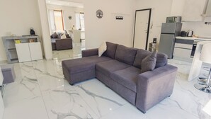 Ein modernes Wohnzimmer mit einem grauen Ecksofa, einem kleinen Couchtisch und einer Küchenzeile inklusive Kühlschrank und Mikrowelle.