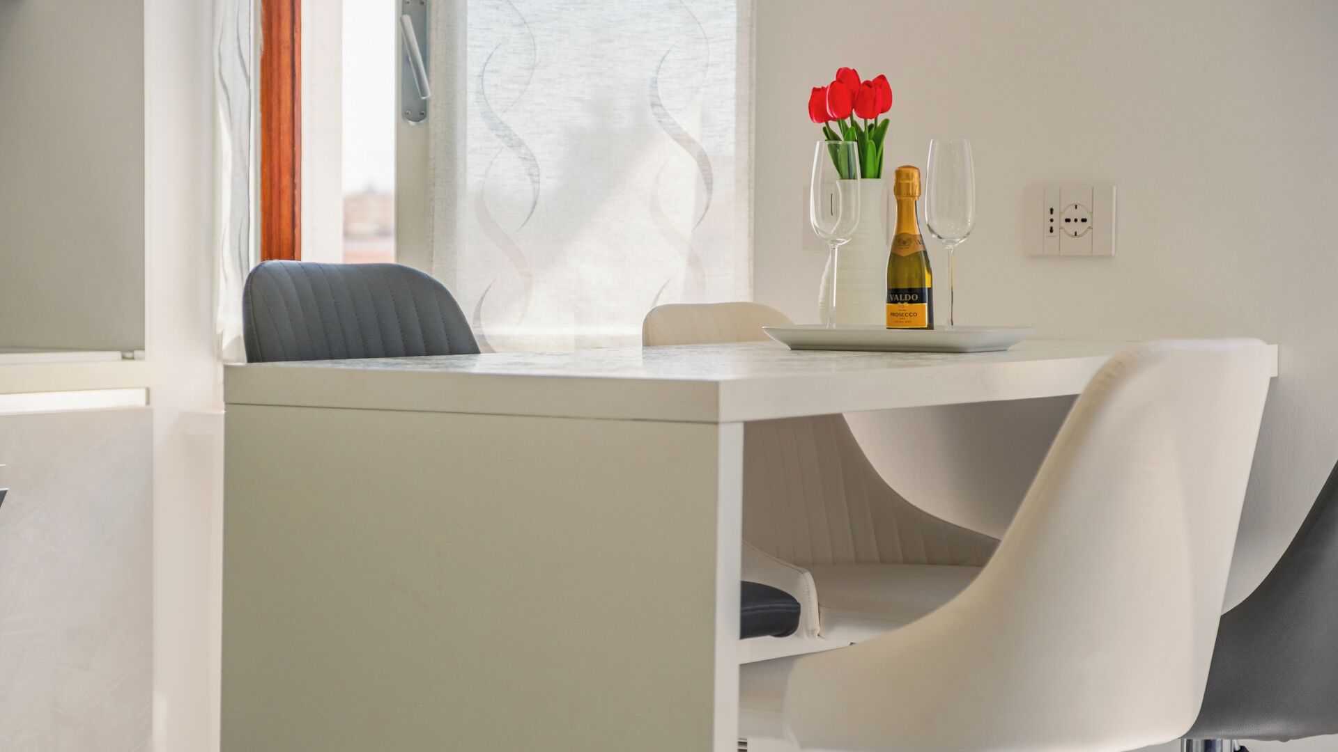 Un coin repas moderne avec une table blanche, deux chaises, une bouteille de vin et un vase rempli de fleurs rouges.