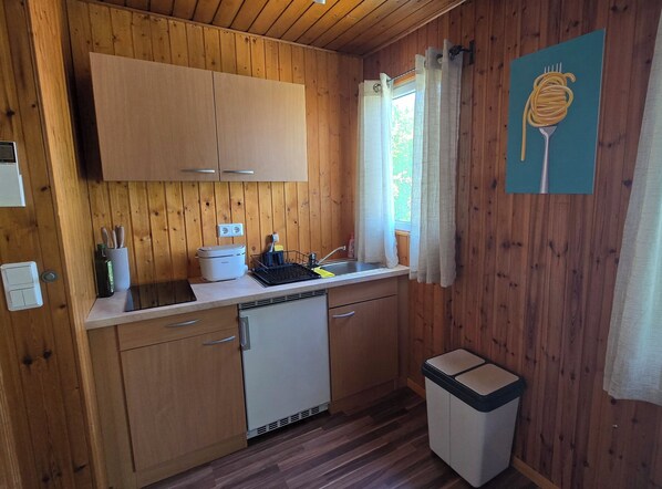 A compact kitchen with wooden walls, a sink, a small refrigerator, and a trash bin. - Cozy Tiny House with a Garden in the Nature Park (Rorodt)