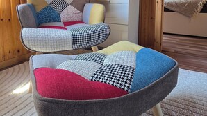 A cozy room with a wooden interior, a comfortable armchair with a patchwork cushion, a small ottoman, and a bookshelf filled with books and decorative items. - Cozy TinyHouse with garden in the nature park (Rorodt)