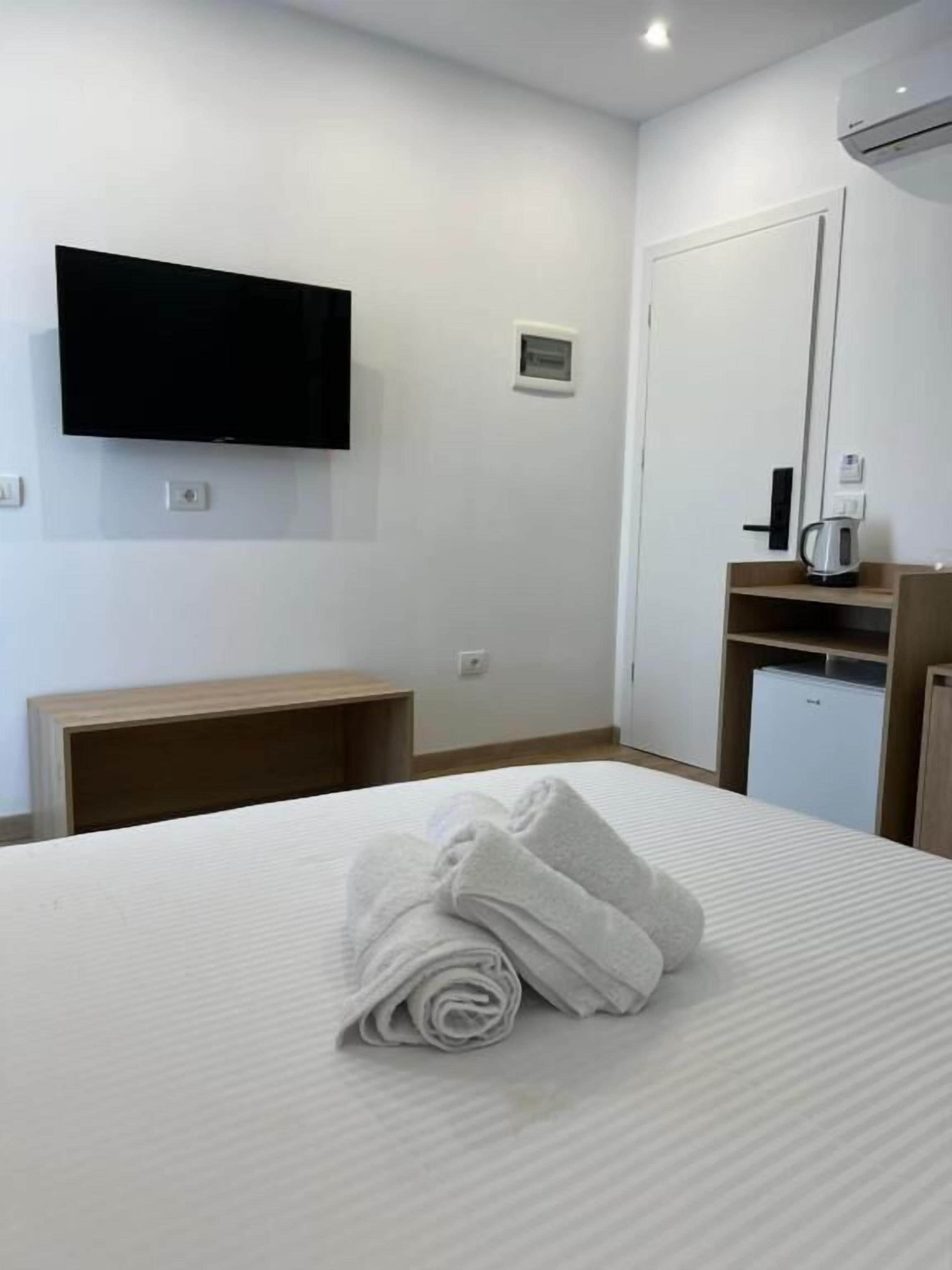 A hotel room with a bed, a flat-screen TV mounted on the wall, a small desk, a coffee maker, and a mini-fridge.