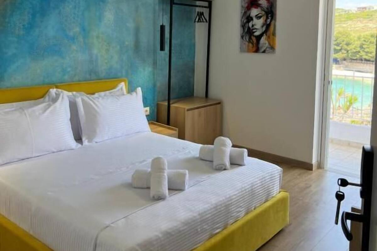 A modern bedroom with a yellow bed, white linens, a blue accent wall, a painting, and a view of a pool.