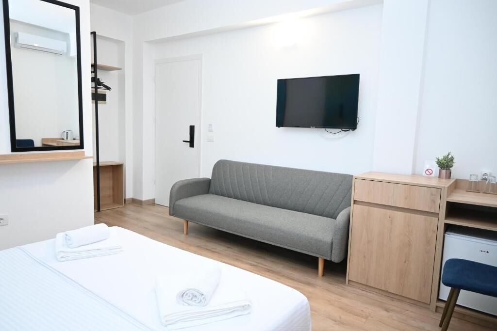 A modern hotel room with a bed, a grey sofa, a TV mounted on the wall, and a small desk with a plant.