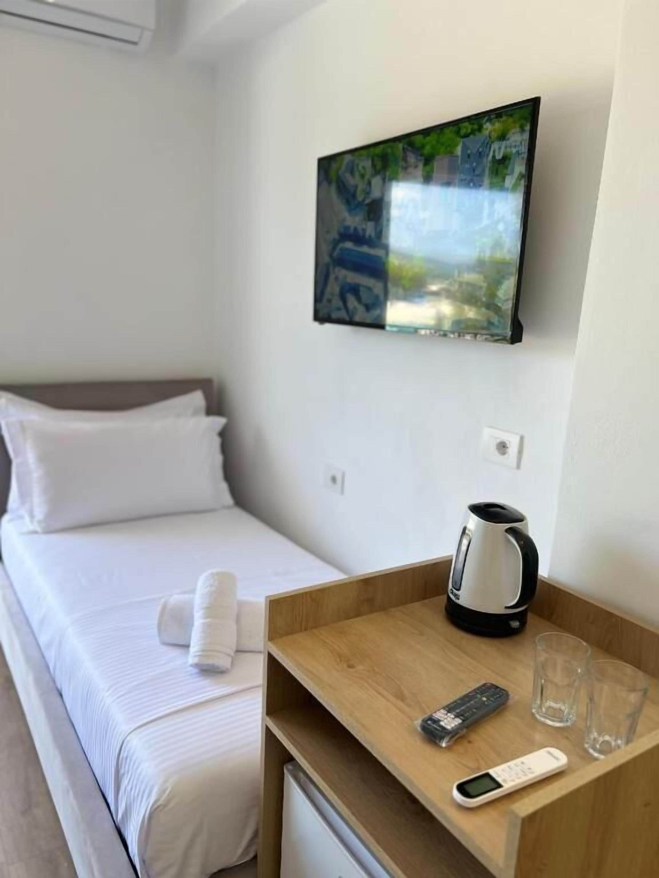 A hotel room with a bed, a TV mounted on the wall, a wooden side table with a kettle, glasses, and a remote control.