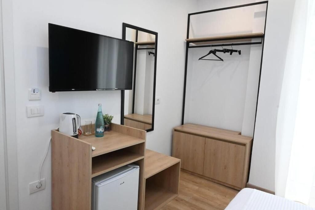 A compact hotel room with a flat-screen TV, a small kitchenette, a wooden wardrobe, and a mirror.