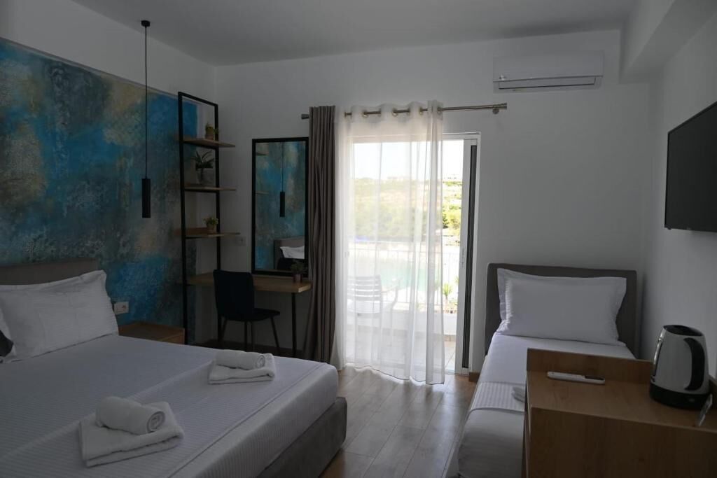 A hotel room with a large bed, a smaller bed, a desk, a chair, and a view of the outdoors through a glass door.