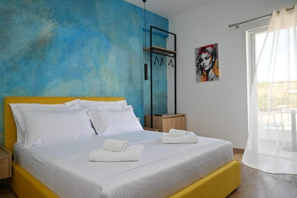 Un dormitorio con cama, ropa de cama blanca, cabecera amarilla, pared azul, un cuadro y una ventana con cortina.