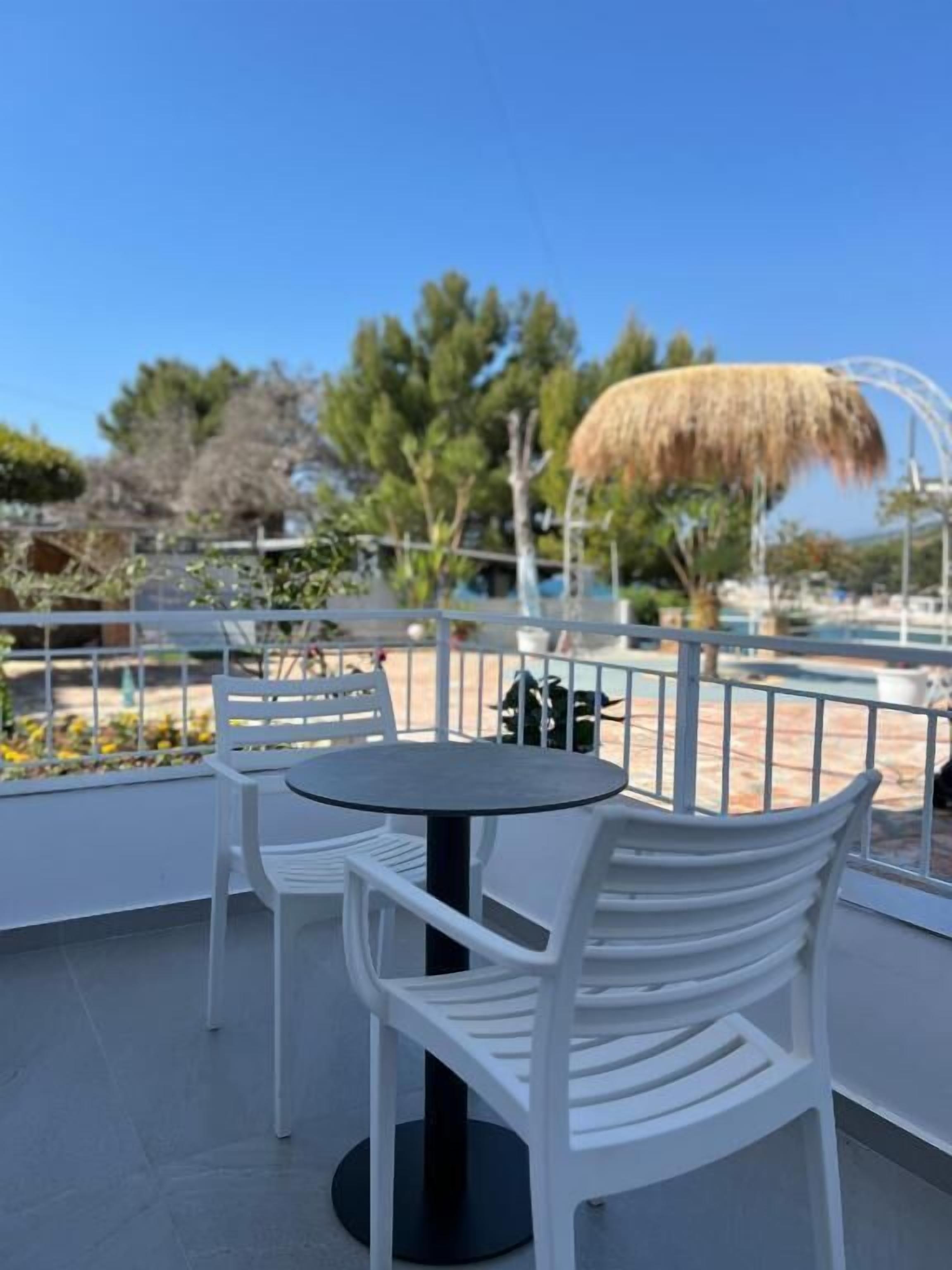 An outdoor seating area with white chairs and a round table, overlooking a pool and trees.