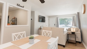 A modern living room with a dining area, a sofa, a TV mounted on the wall, and a ceiling fan. - Cozy Pet Friendly 3BR - Fenced Yard - 1Mi HPU & DwnTwn - Furniture Market (High Point)