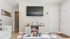 A modern living room with a flat-screen TV mounted on the wall, a white sofa, a coffee table with remote controls, a cabinet with decorative items, and a door with a doormat. - Cozy Pet Friendly 3BR - Fenced Yard - 1Mi HPU & DwnTwn - Furniture Market (High Point)