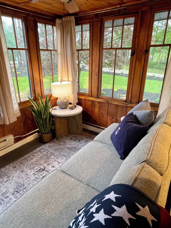 A cozy room with a wooden ceiling, a window with a view of a garden, a round table with a lamp, and a sofa with patterned cushions.