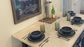 A dining area with a long table set for four, featuring black plates, clear glasses, and a vase with white flowers. A framed fish artwork hangs above the table.