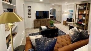 A modern living room with a brown sofa, a coffee table, and a TV. There are bookshelves, a lamp, and a dining area in the background.