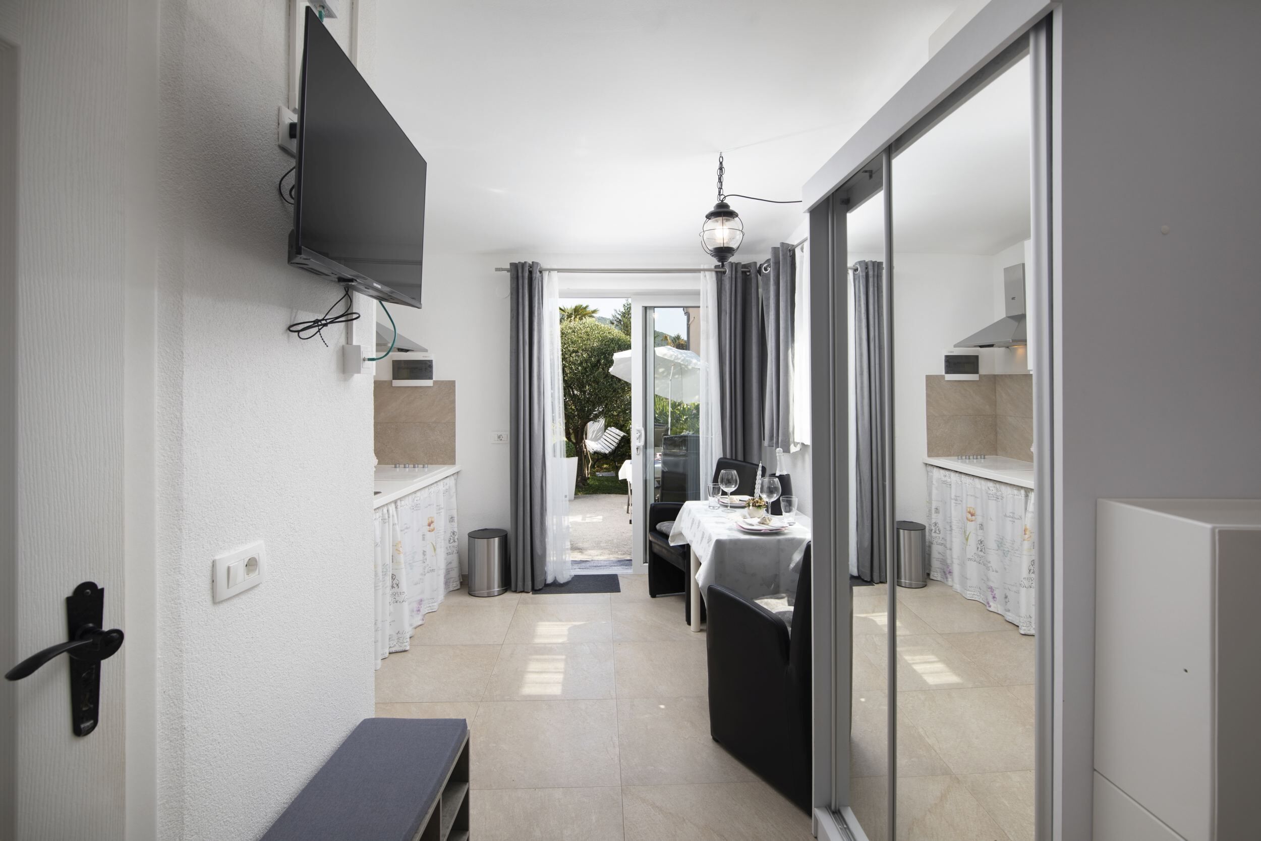 A modern hotel room with a flat-screen TV mounted on the wall, a dining area with a table set for two, and a view of the outdoor garden through the sliding glass doors.