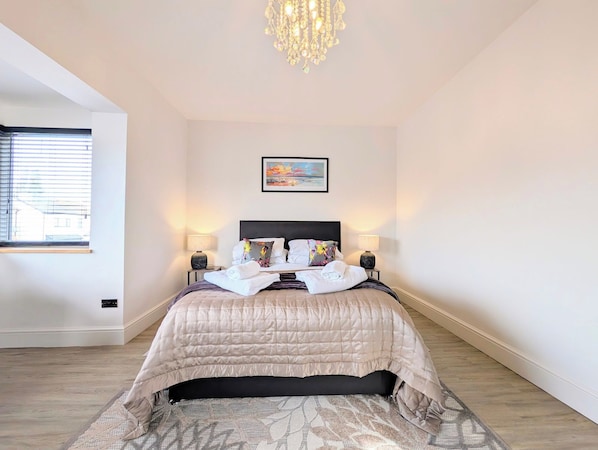 A hotel room with a bed, two bedside lamps, a chandelier, a window with blinds, and a painting on the wall.