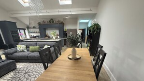 A modern living room with a wooden dining table, grey sofa, and black chairs. A chandelier hangs from the ceiling, and there is a kitchen area in the background.