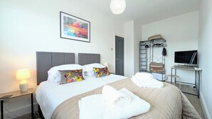 Quarto de hotel com cama, escrivaninha, televisão, guarda-roupa e um quadro na parede.