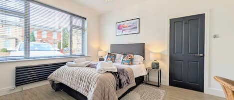A bedroom with a bed, bedside lamps, a window with blinds, a door, and a chandelier.