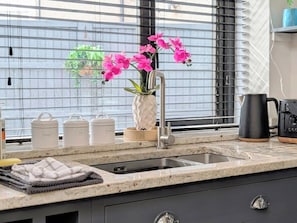 A kitchen with a sink, a window with blinds, a vase with flowers, and kitchenware.