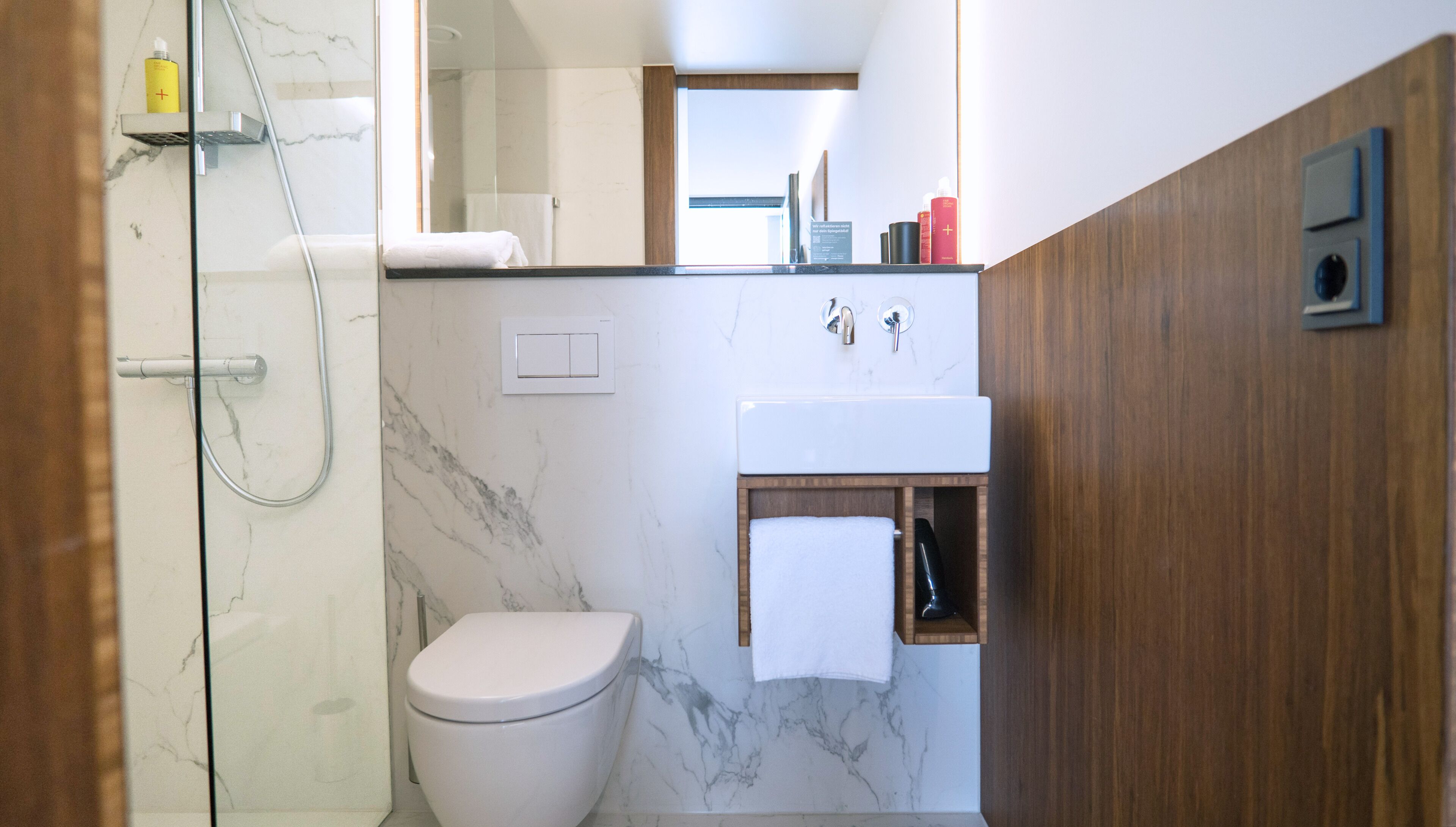 A modern bathroom with a glass shower enclosure, a white toilet, a wooden vanity with a mirror, and a marble backsplash.