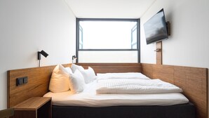 Una habitación de hotel moderna con cabecera de madera, un ventanal grande, un televisor de pantalla plana montado en la pared y una mesita de noche con lámpara.