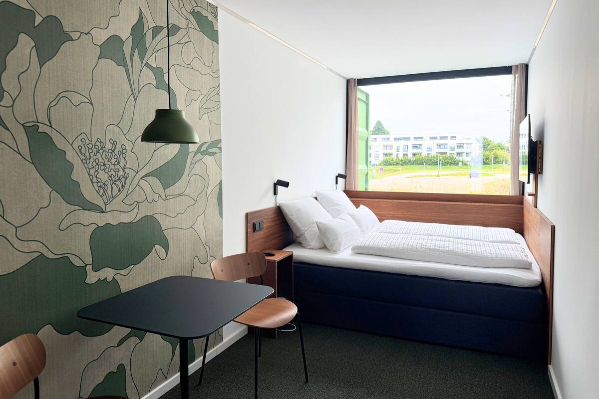 Camera d'albergo moderna con un letto, una scrivania, una sedia e un'ampia finestra che si affaccia su edifici e vegetazione.