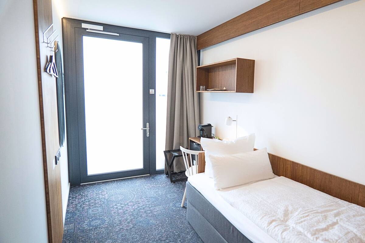 Una camera d'albergo con un letto, una scrivania con una sedia, un piccolo ripiano e una porta che dà su un balcone.