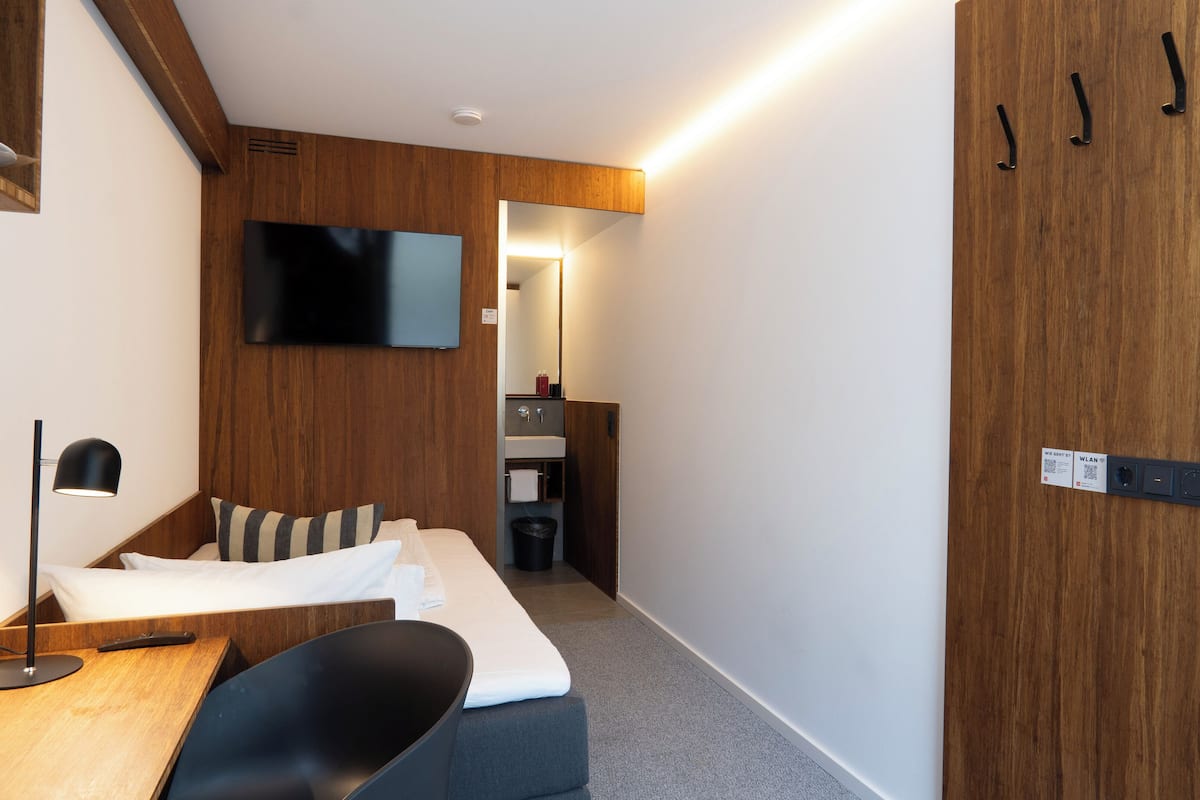 Una camera d'albergo moderna con una parete in legno decorativa, una TV a schermo piatto sopra il letto, una scrivania con sedia e un piccolo bagno con lavandino e specchio.
