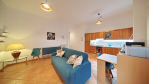 A modern living room with a blue sofa, a glass coffee table, and a dining area with a small table and chairs. A kitchen is visible in the background with wooden cabinets and a countertop.