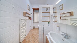 A bathroom with white tiled walls, a white sink, and a shower area. There are wooden shelves above the sink and a towel rack on the wall.