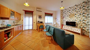 A modern kitchen with wooden cabinets, a dining area with a table and chairs, a sofa, and a TV on the wall.